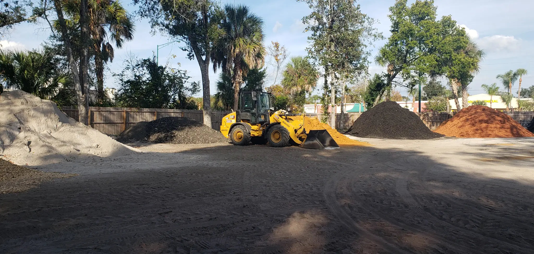 Mulch and Stuff landscape materials yard in Ormond Beach, showing CAT loader and stocked piles of mulch, sand, and aggregates for pickup and delivery.